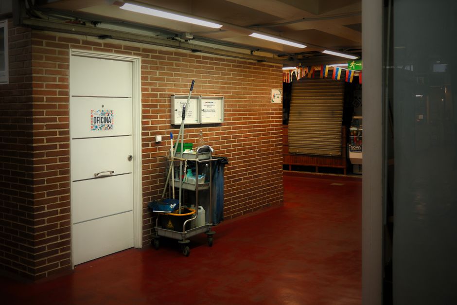 Two professional cleaners are performing window cleaning at the entrance of a commercial building, using long-handled squeegees to wash the large glass doors and panels. The interior features tiled fl